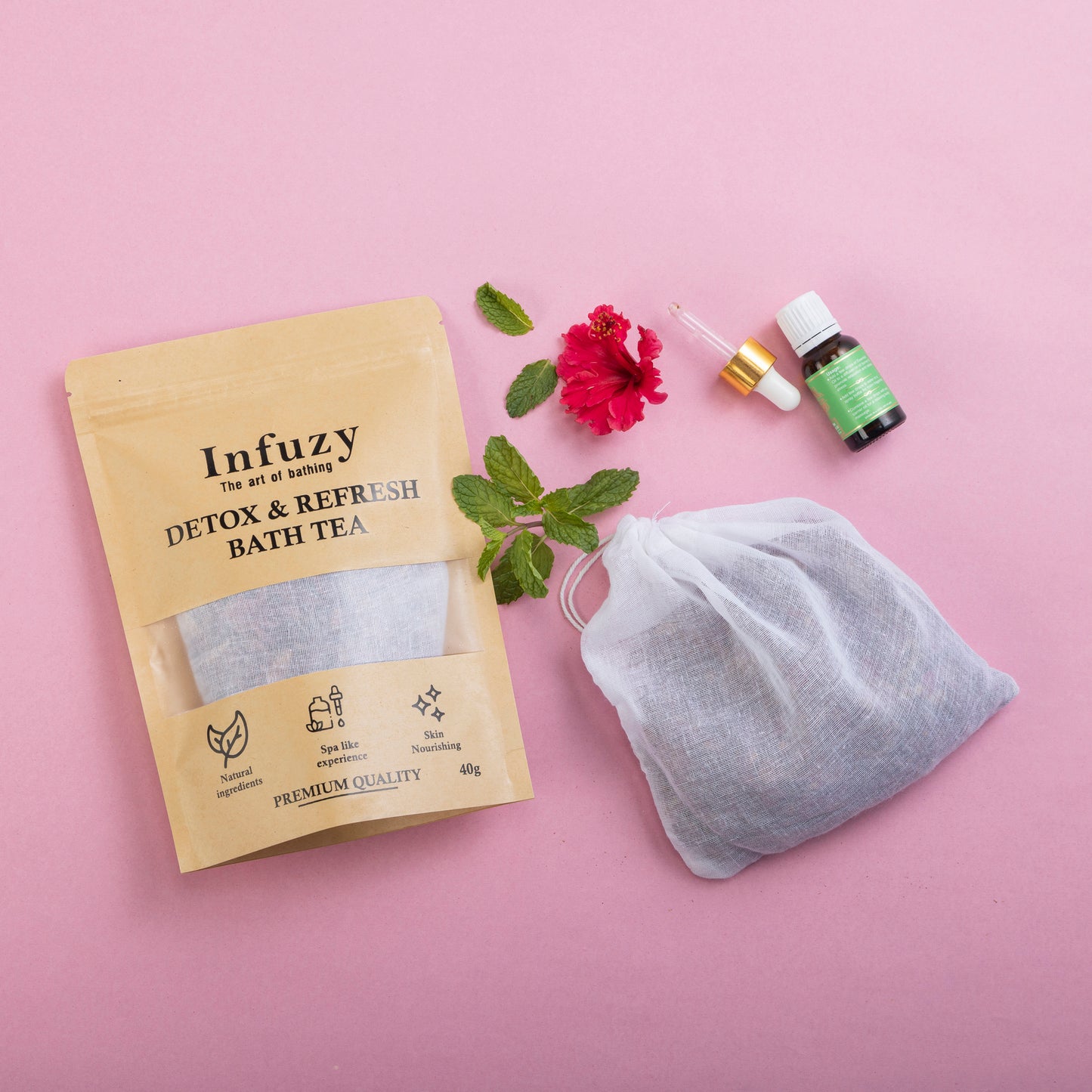 A package of Infuzy Detox & Refresh Bath Tea with a tea bag, green and red flowers, and a bottle of essential oil displayed on a pink background.