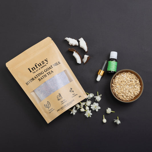 A package of Hydrating Goat Milk Bath Tea by Infuzy, with ingredients like goat milk powder, jasmine petals, and a vial of vitamin E oil displayed on a dark background, along with a small bowl of oats and a candle.